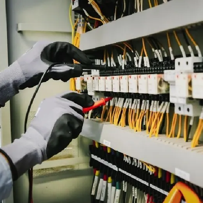 Gloved hands testing electrical wiring in a panel with rows of switches and yellow cables