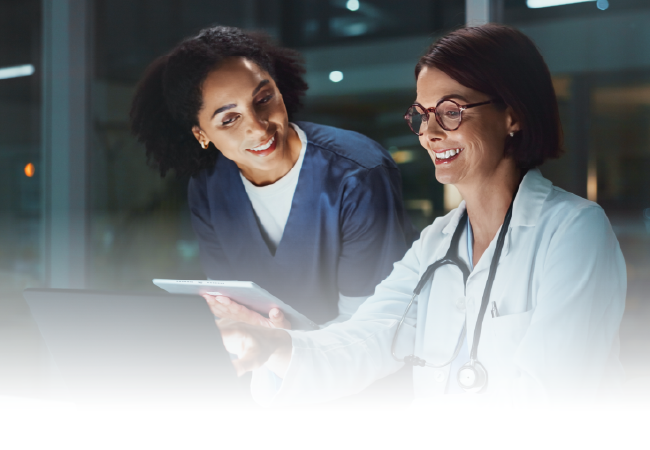 Two coworkers smiling over a tablet in a dim office, with one wearing a stethoscope