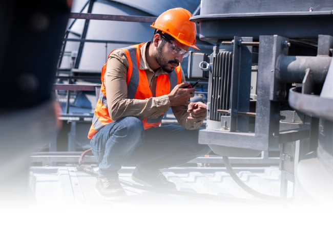 Worker in orange hard hat kneeling to inspect industrial machinery in a factory.