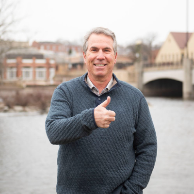 Smiling man in a gray sweater giving a thumbs-up by a river with buildings in the background
