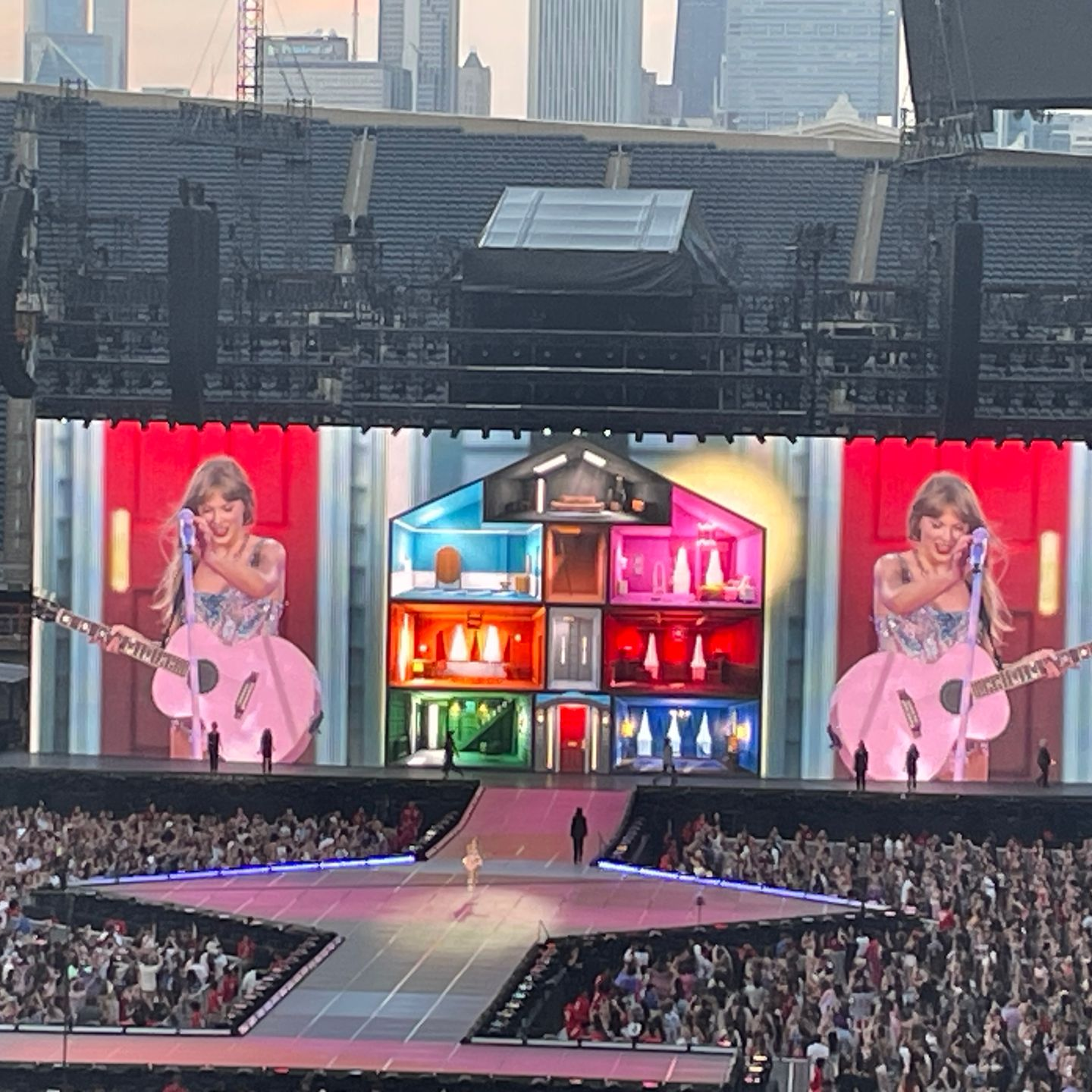 Concert stage with giant screens, colorful house set, and crowd in a stadium at dusk
