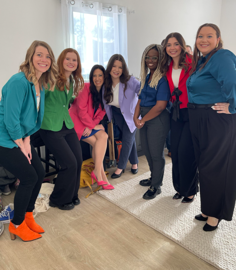 Six women smiling and posing indoors in colorful outfits near a bright window.