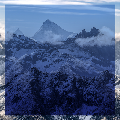 Snow-covered mountain peaks under a cloudy blue sky, with a dark forested ridge in the foreground
