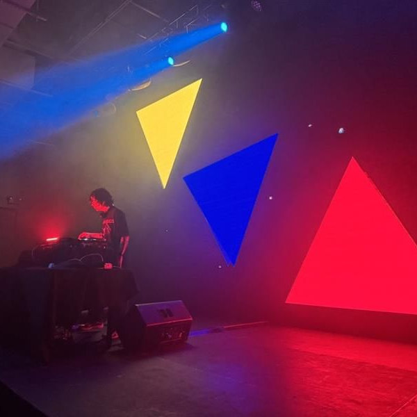 DJ performing on a foggy stage with large yellow, blue, and red triangular lights