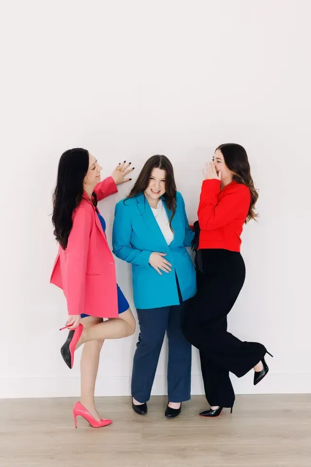 Three people posing in bright blazers against a white wall, one holding a cup and the others laughing.