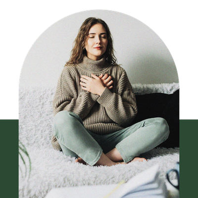 Woman sitting cross-legged on a bed, hands over heart, wearing a beige sweater and green pants.