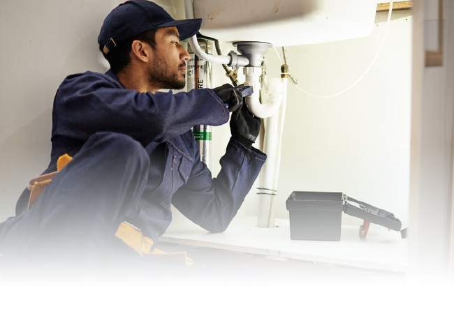 Technician wearing gloves and cap repairs machinery indoors, using a tool near a white ceiling unit.