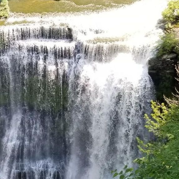 Wide waterfall cascading over a rocky cliff, surrounded by green foliage and bright sunlight.