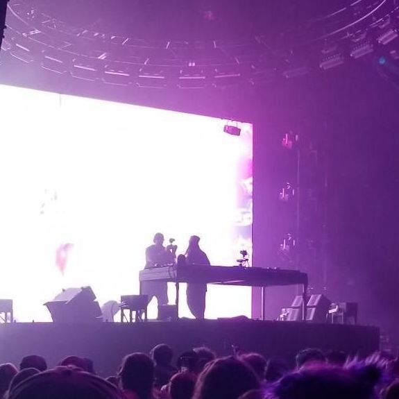 Concert stage with bright white screen, purple lighting, and silhouetted DJ performers above a crowd