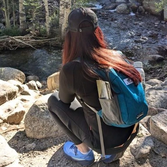 Person crouching on rocks beside a creek, wearing a blue backpack and bright blue shoes.