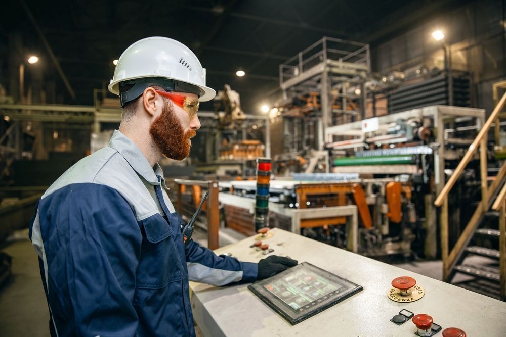 Worker in hard hat operating a control panel on a factory floor with machinery in the background