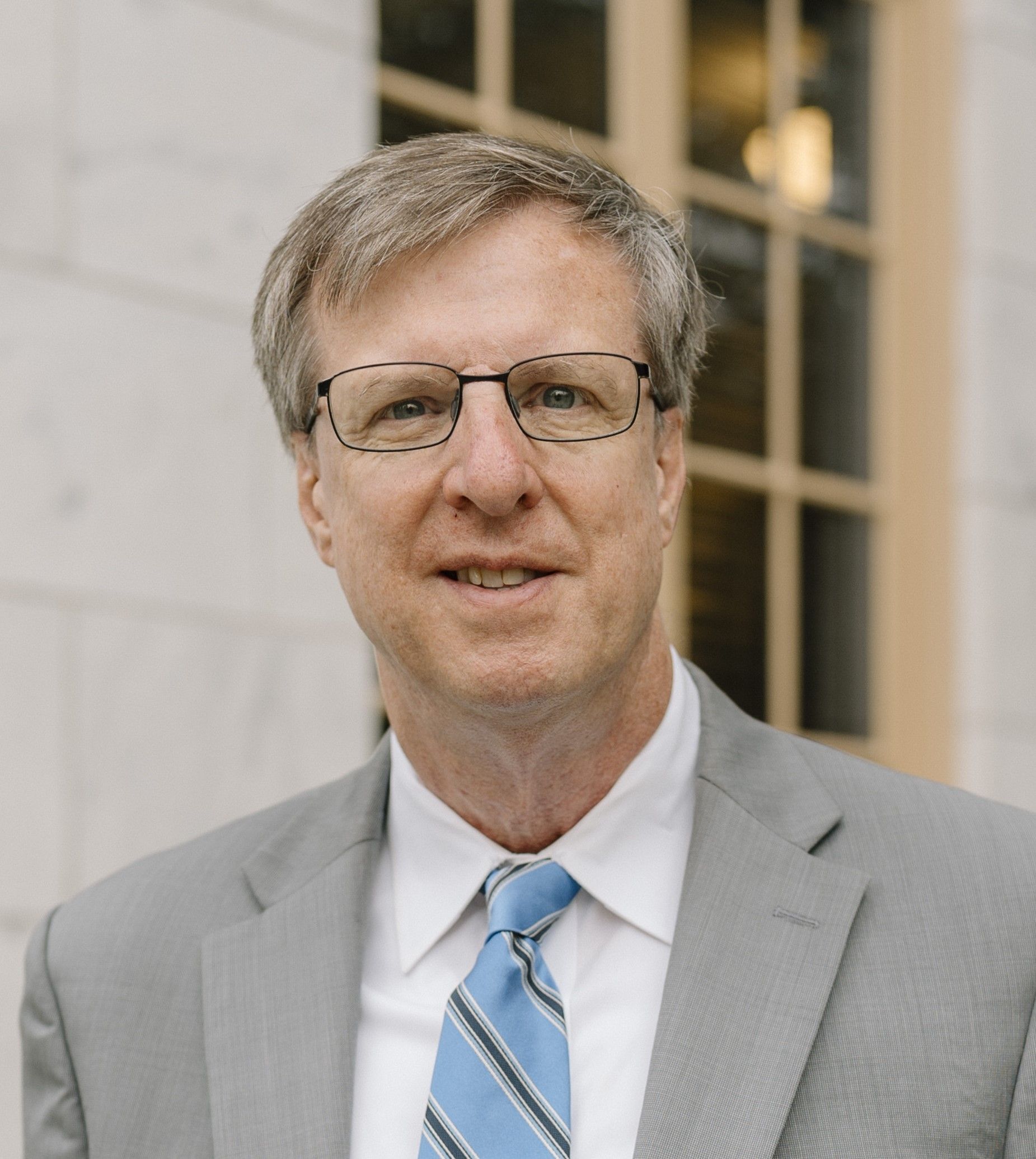 Man in suit and tie wearing glasses, standing outside a building, smiling.