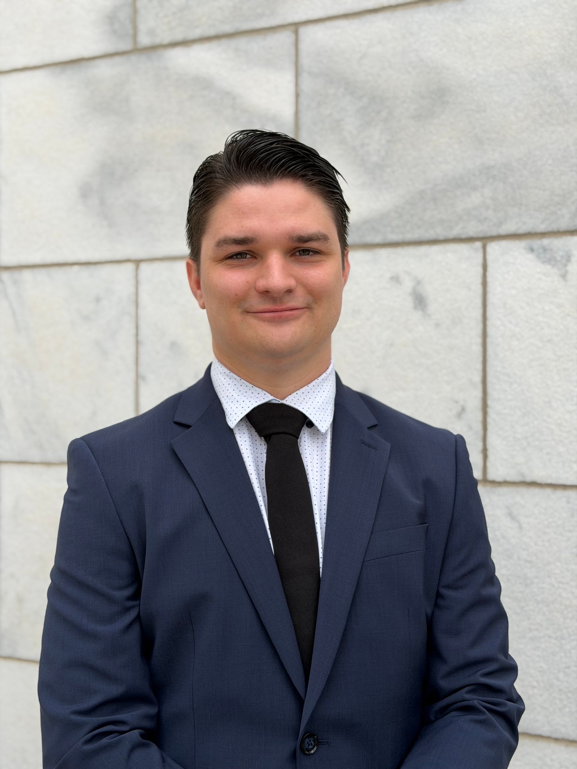 Man in a suit and tie, posing in front of a light-colored building.