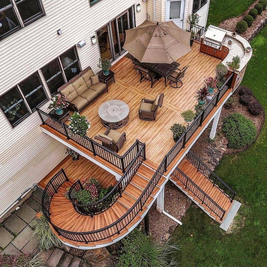 Multi-level wooden deck with curved stairs, outdoor furniture, and an umbrella, extending from a house to a grassy yard.