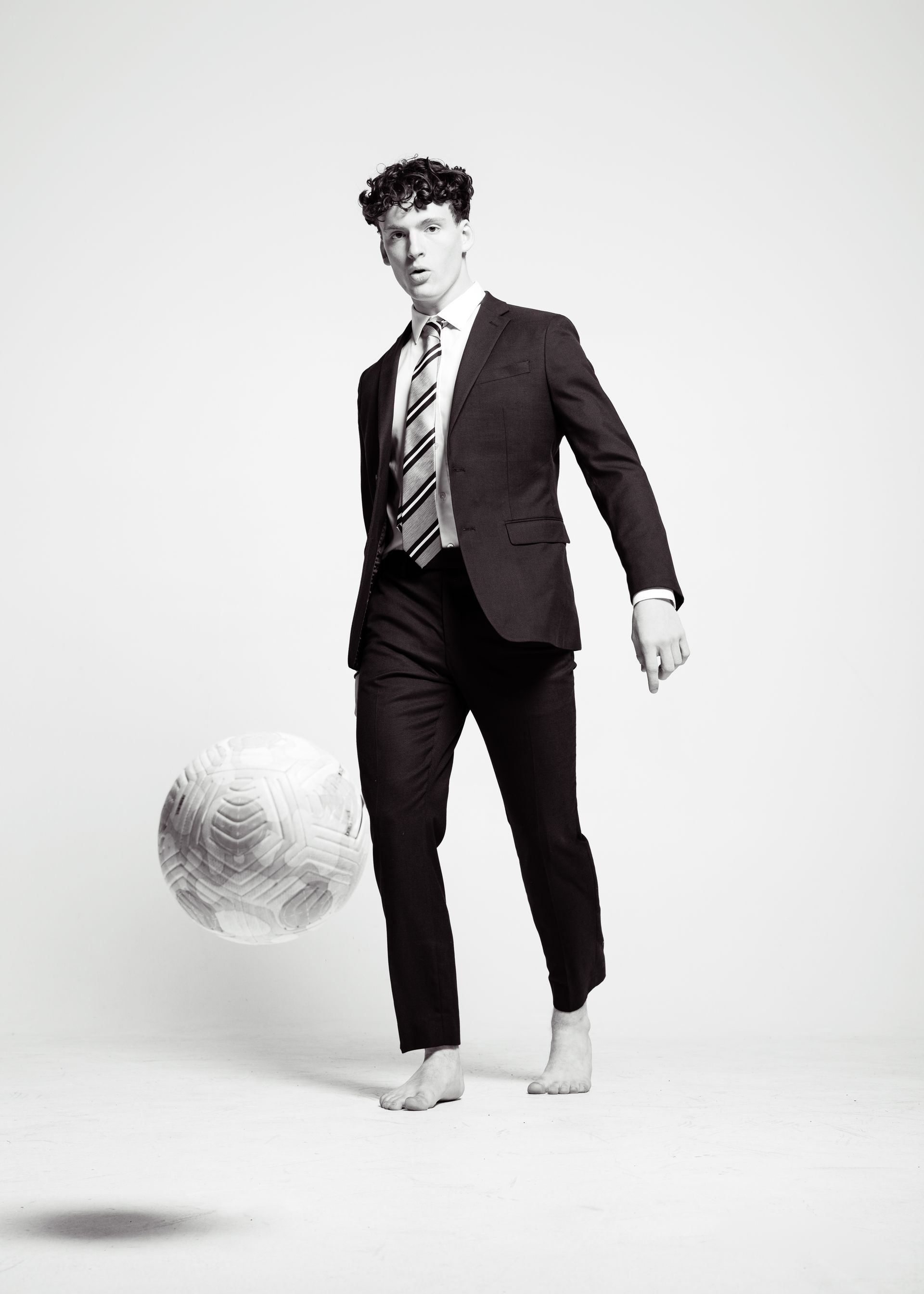 Man in suit barefoot, juggling a sphere against a white background.
