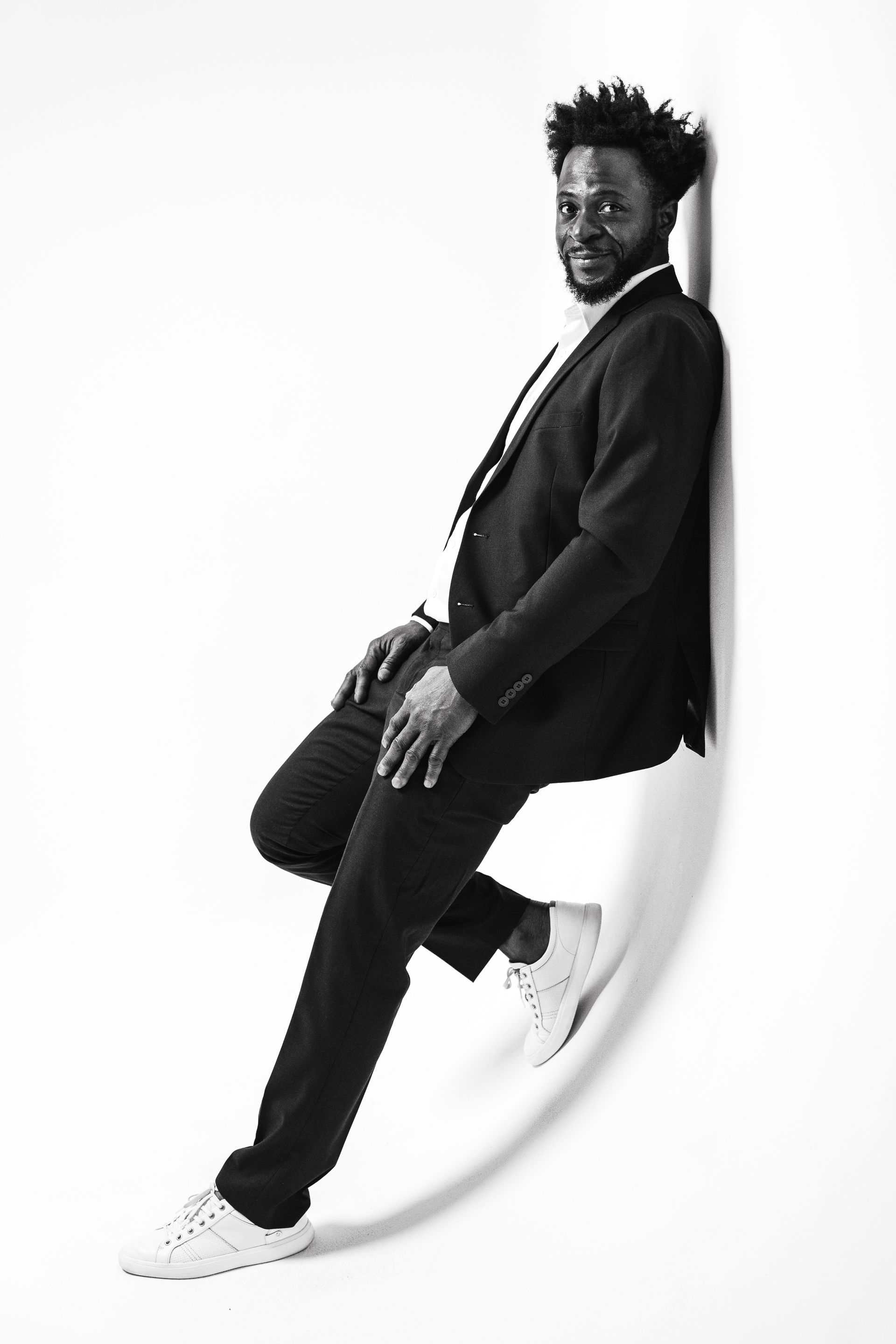 Man in suit and sneakers leans against a white wall, smiling.