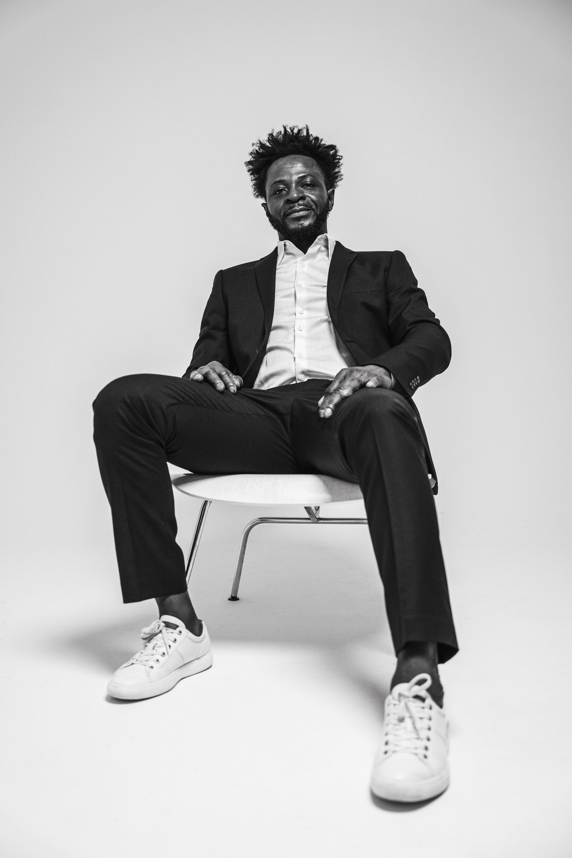 Man in suit and sneakers sits on a chair, smiling at the camera, in a studio.