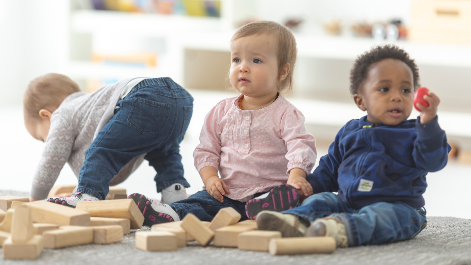 Three toddlers playing with wooden blocks on a carpet, one bending over, two sitting nearby.