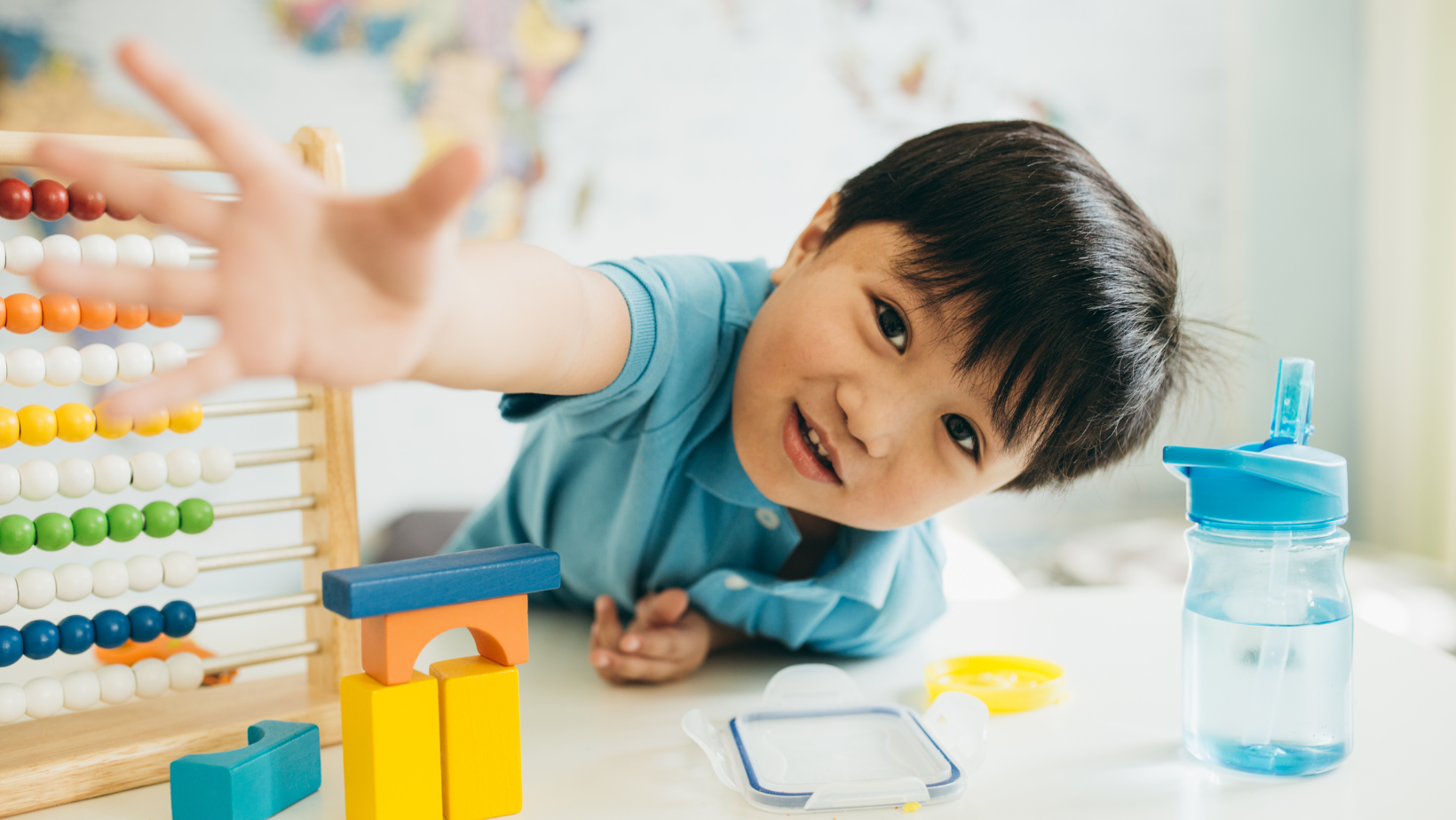 Child reaching across a table with an abacus and water bottle, smiling in a bright classroom