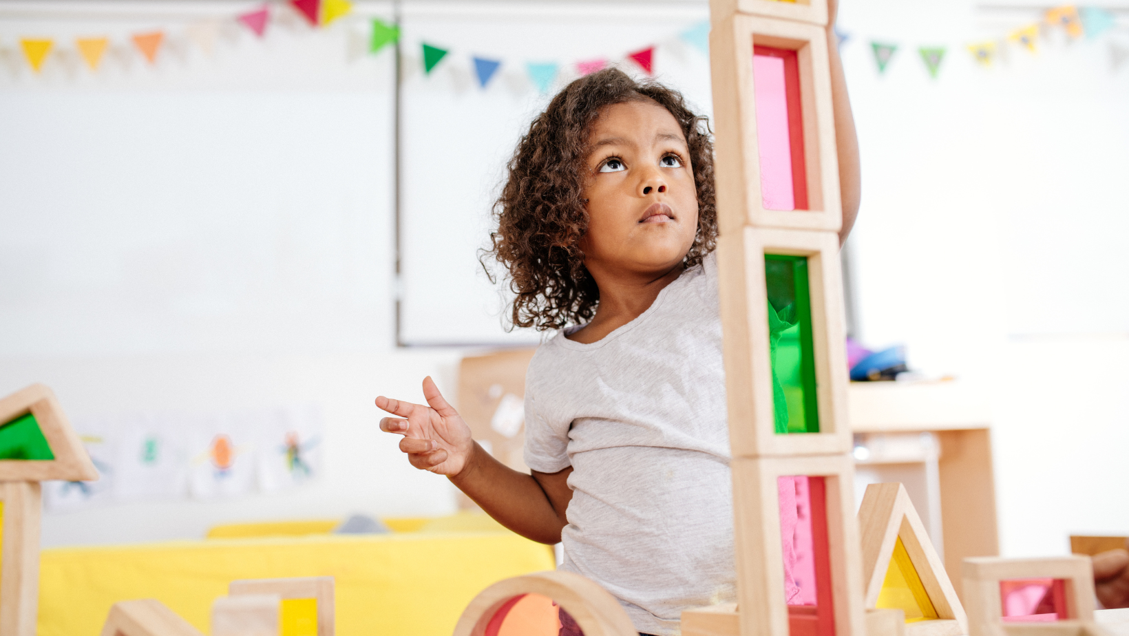 Child playing with colorful wooden blocks in a bright playroom, looking up thoughtfully.