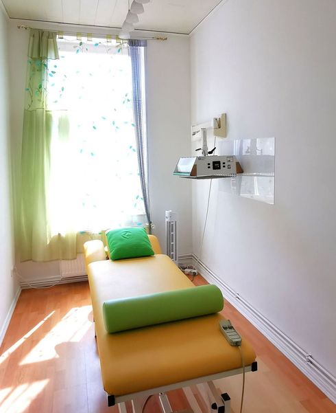 Von der Sonne beschienene Massageliege in einem hellen Behandlungsraum mit grünem Kissen und wandmontierter Ausstattung