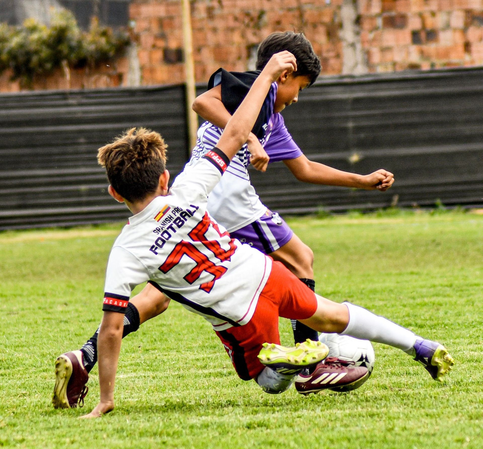 Two soccer players collide while tackling on a grassy field, one in white and red, the other in purple.