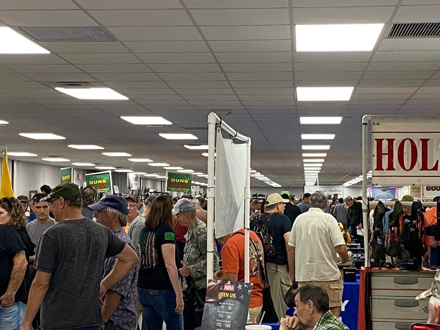 Crowd browsing at an indoor fair, numerous booths and displays, bright overhead lighting.