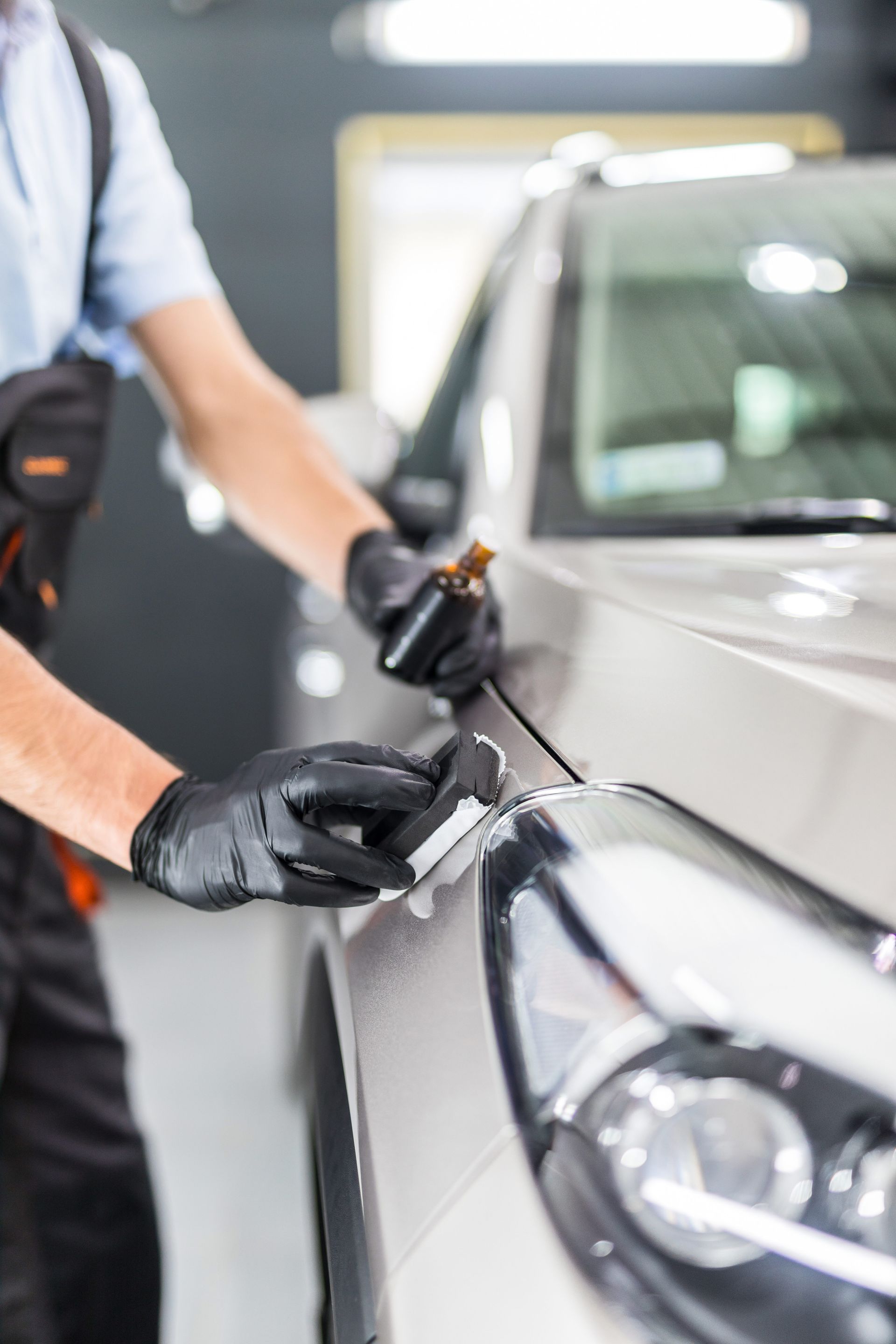 A person in black gloves applying a coating to a car's hood, likely for detailing or protection, in a well-lit garage.