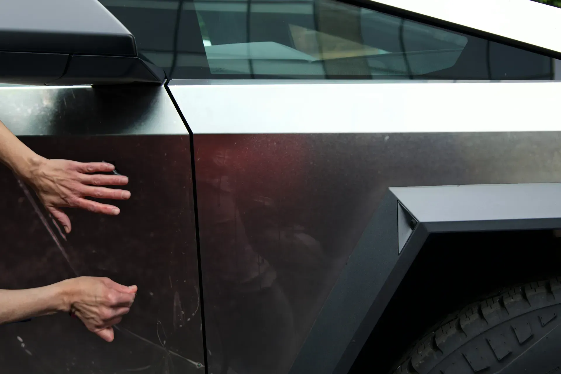 Hands touching the scratched metal panel of a Tesla Cybertruck.  The metallic surface reflects a blurry background.