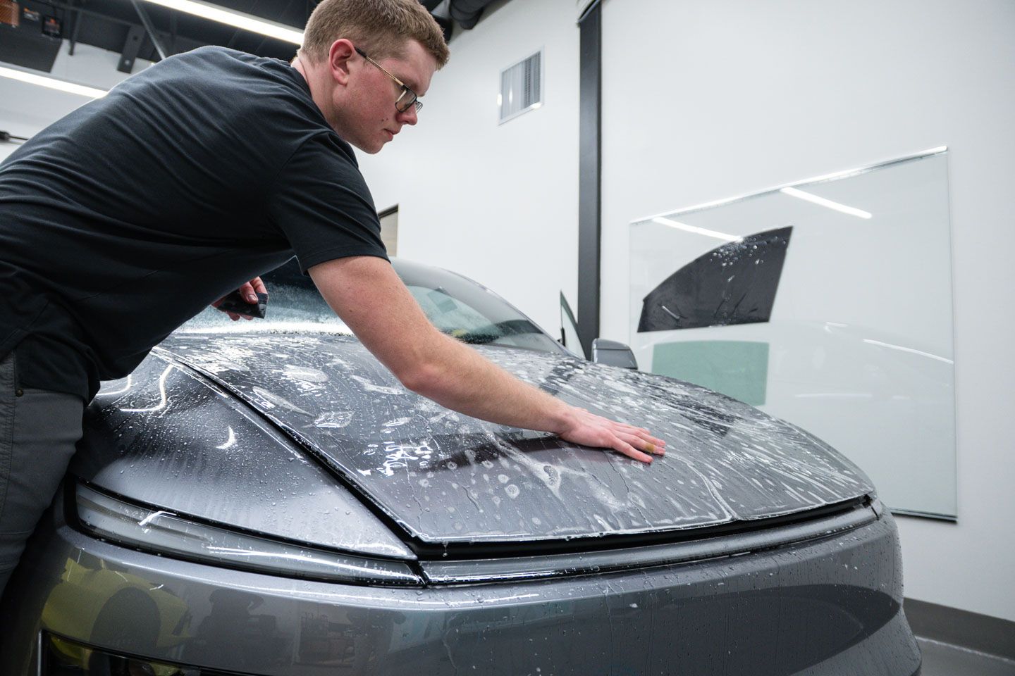 A man applies protective film to the hood of a gray car inside a bright shop. He wears glasses and a black shirt.