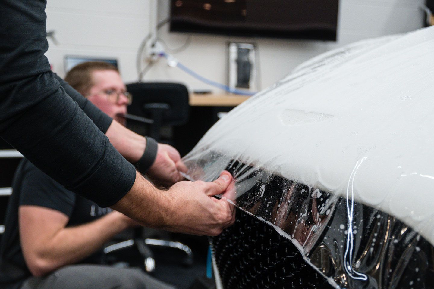 Hands applying clear protective film to a white car hood in a workshop