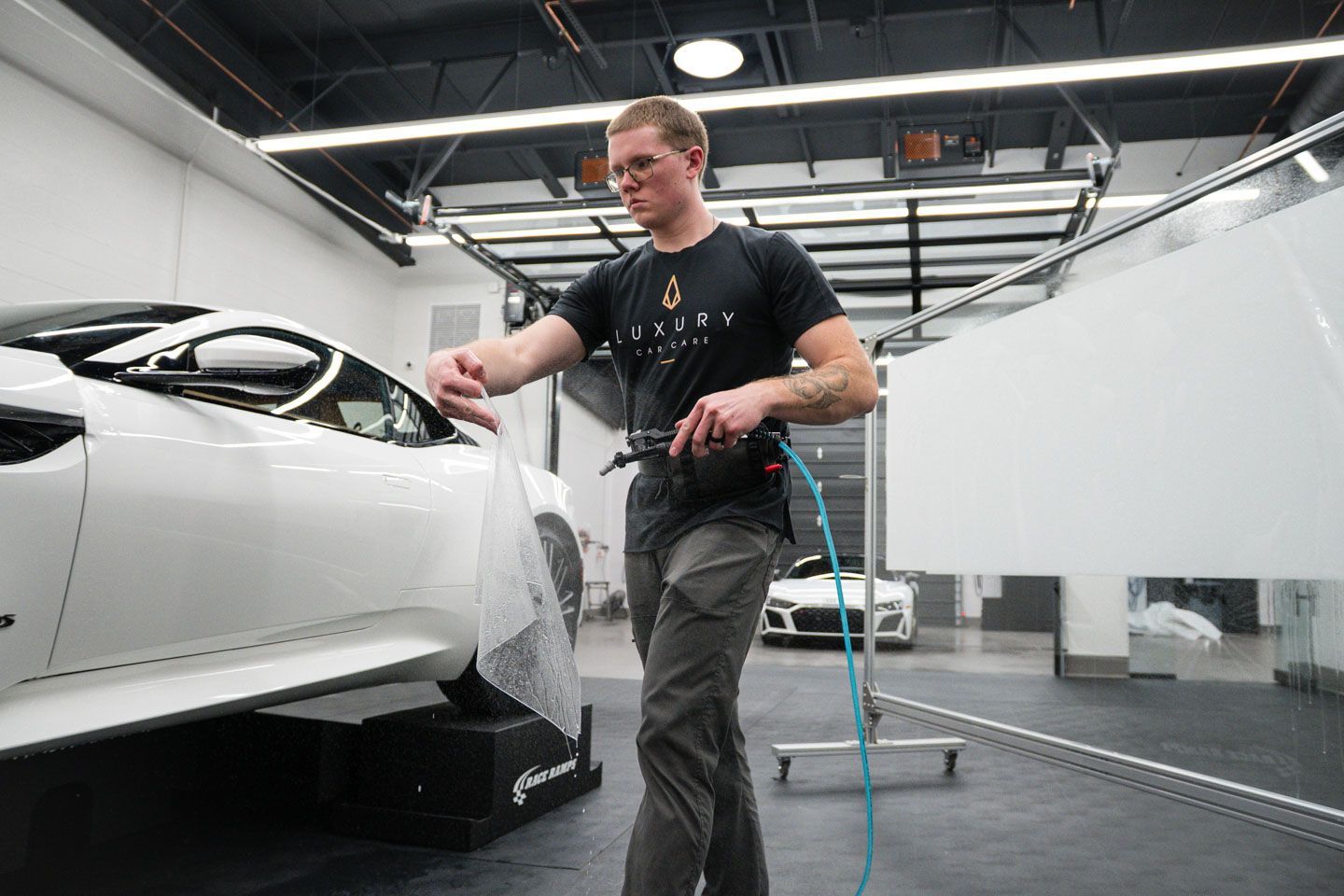 A man applies a film to a white car in a workshop. He holds a tool, likely for smoothing the film.