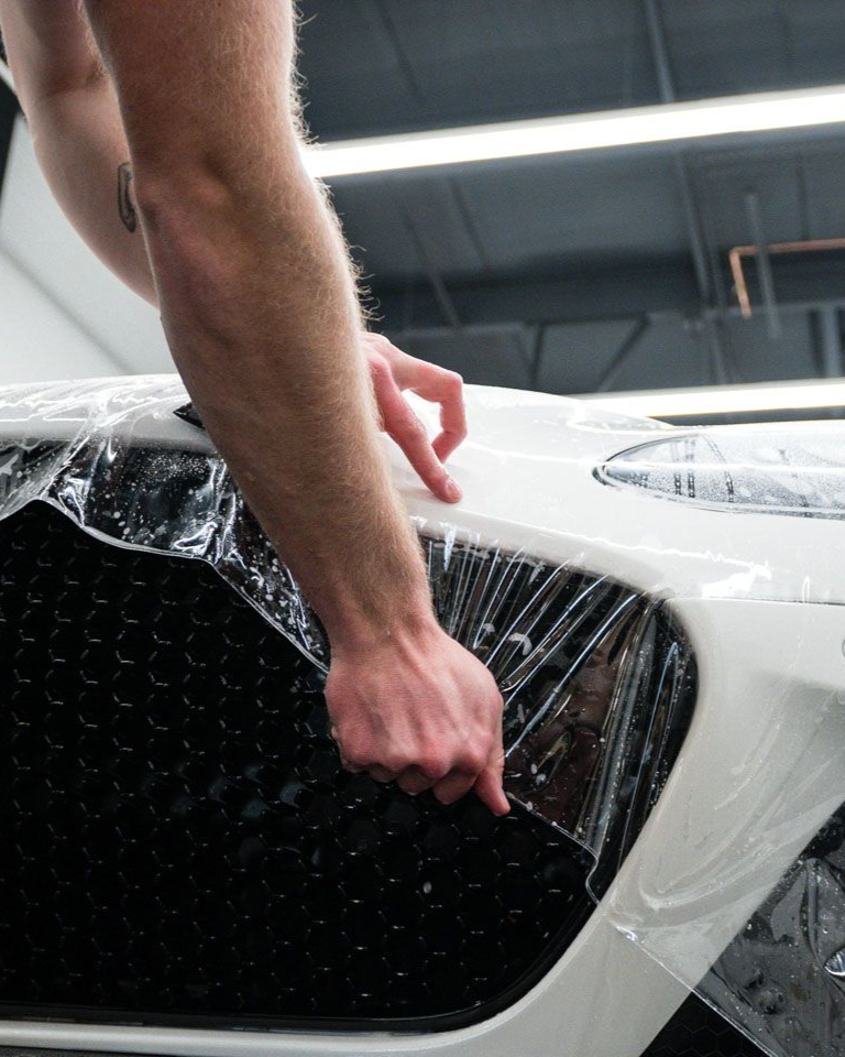 Person in black gloves applying sealant to a black car's hood in a well-lit garage.