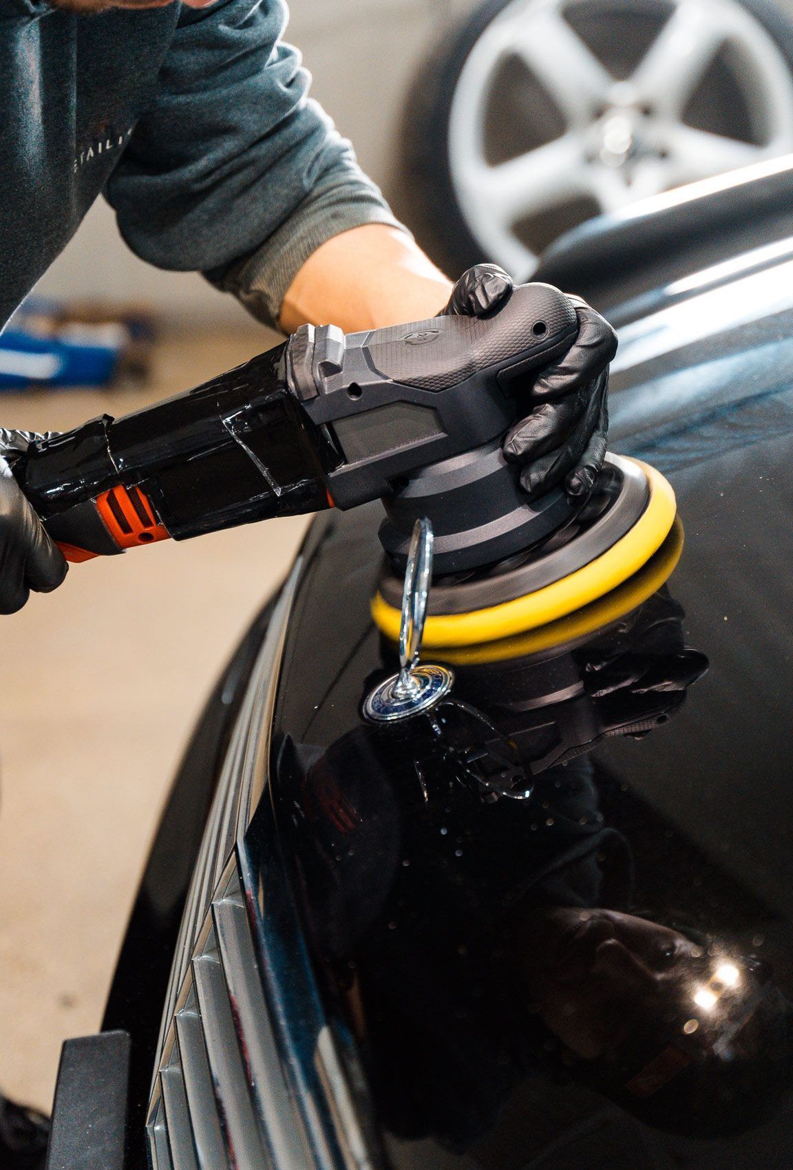A person wearing black gloves polishes a black car with a power buffer in a garage.