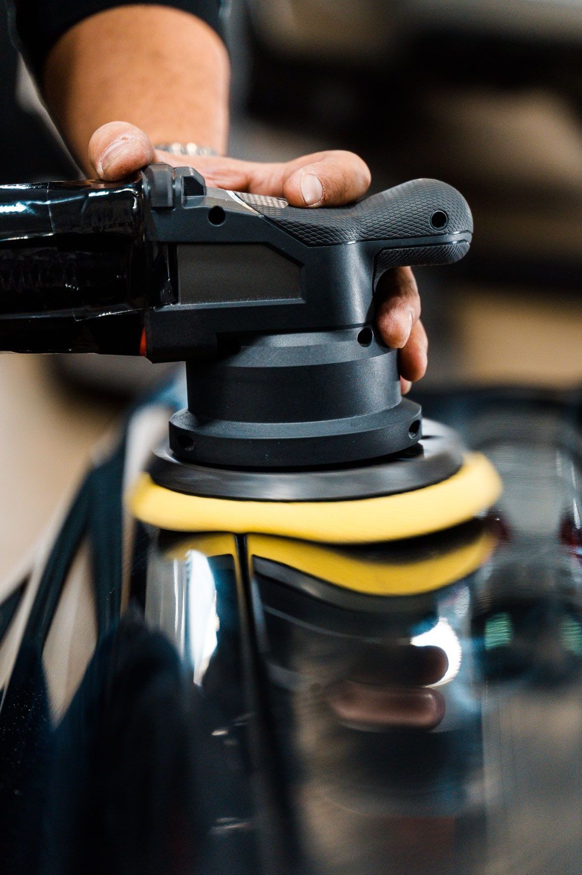 A person polishing a black car door with a black and yellow buffer in a garage.