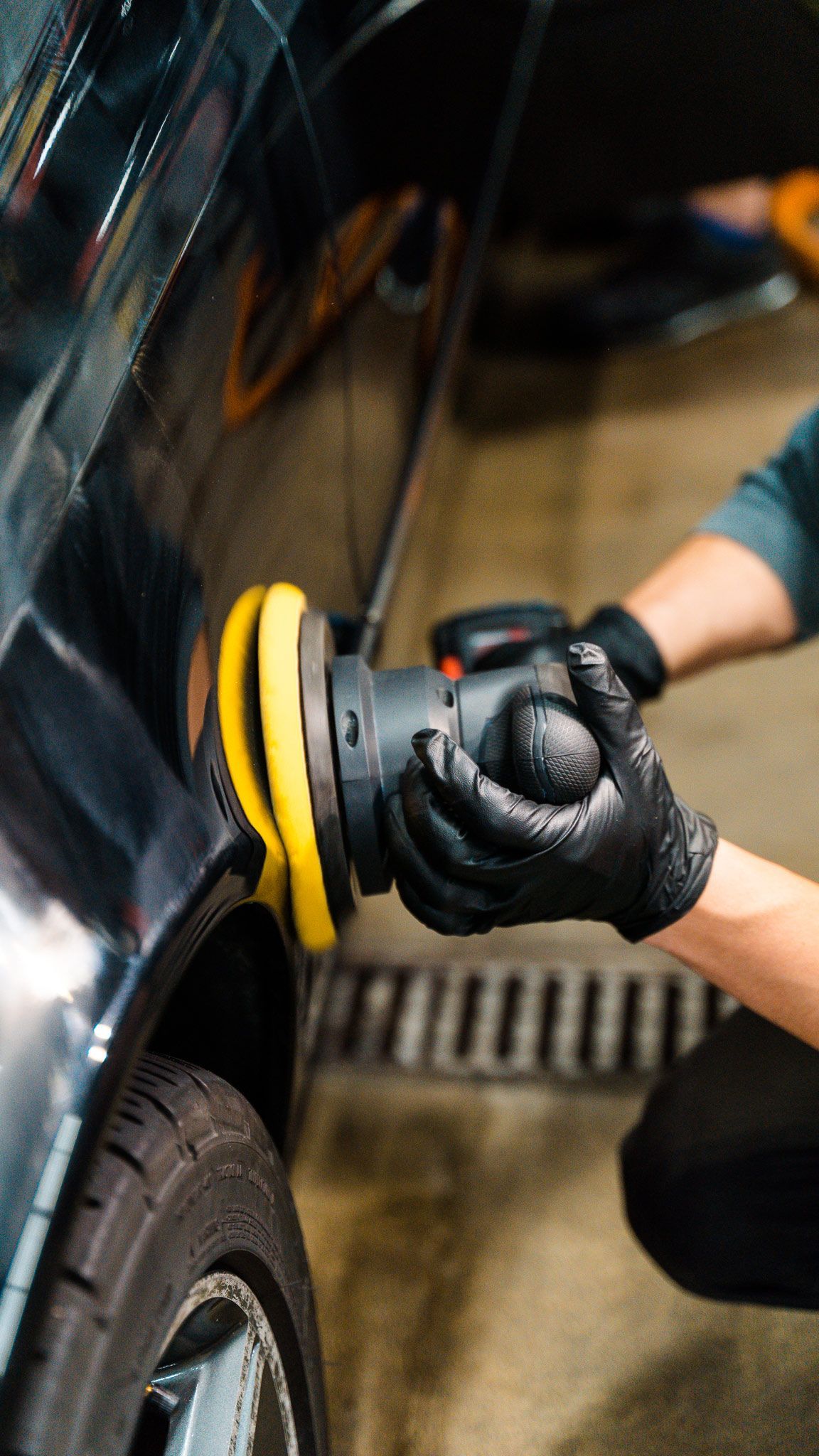 A person wearing black gloves polishes a black car with a yellow and black buffer in a garage.