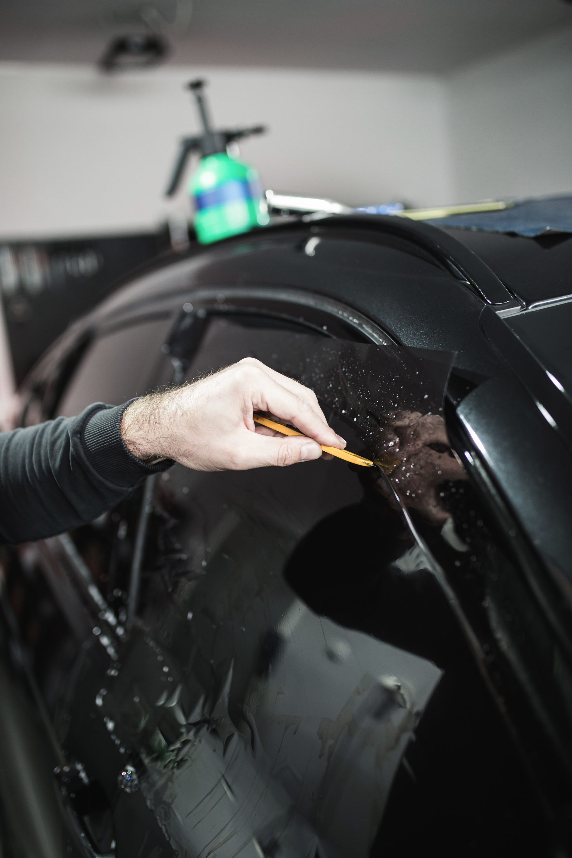 A person in a tan cap applies tinted film to a car window, indoors. They use their hands to smooth the film against the glass.