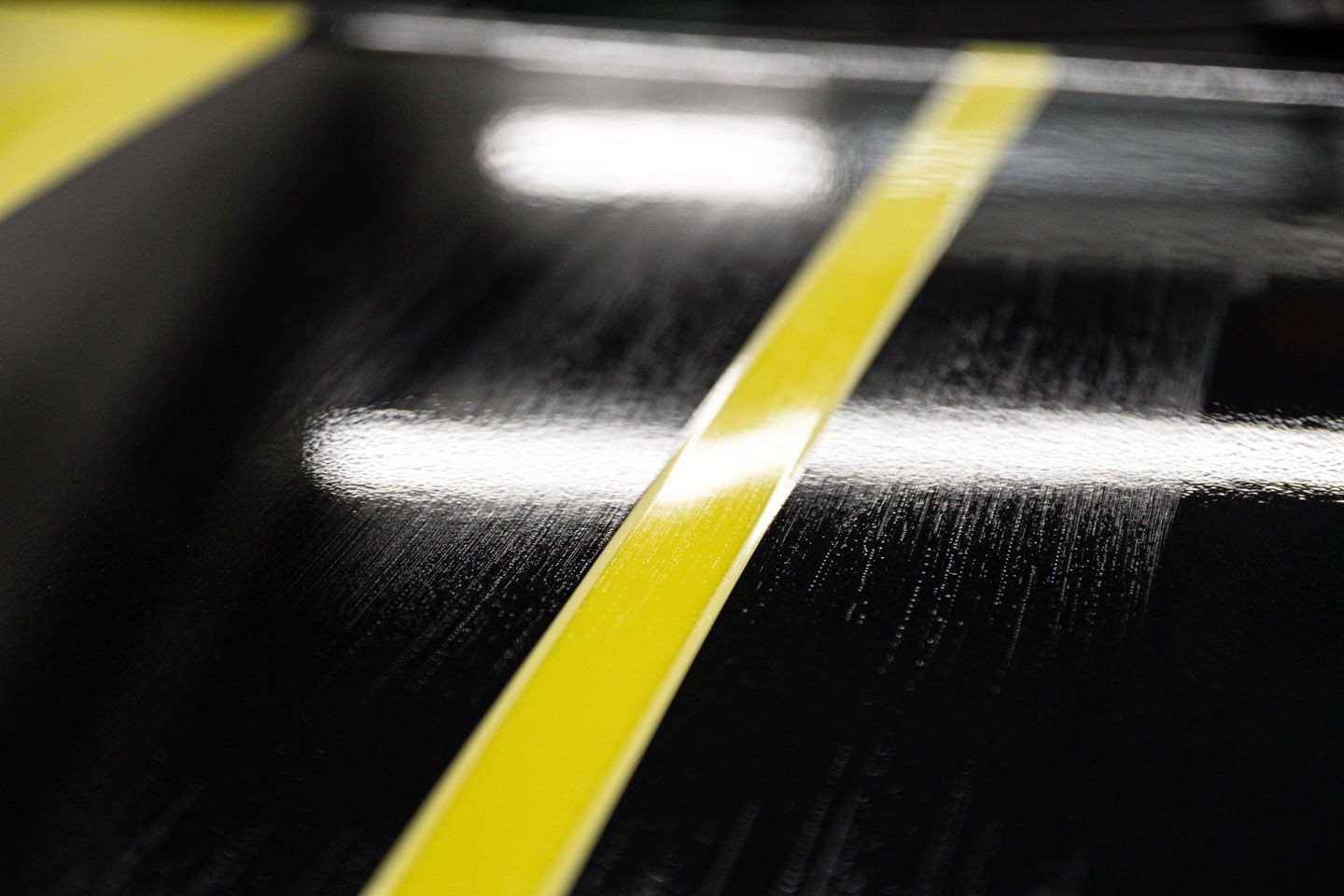A black surface with a bright yellow tape stripe, possibly for detailing, with visible reflections.