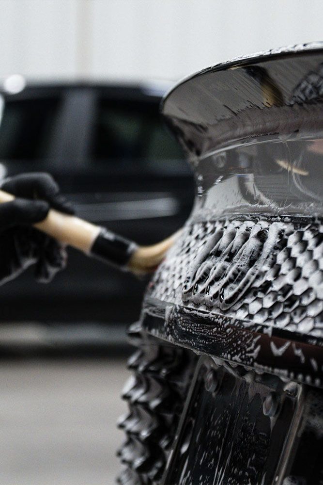 A person with a gloved hand uses a brush to clean a black car spoiler covered in soap. A black vehicle is in the background.