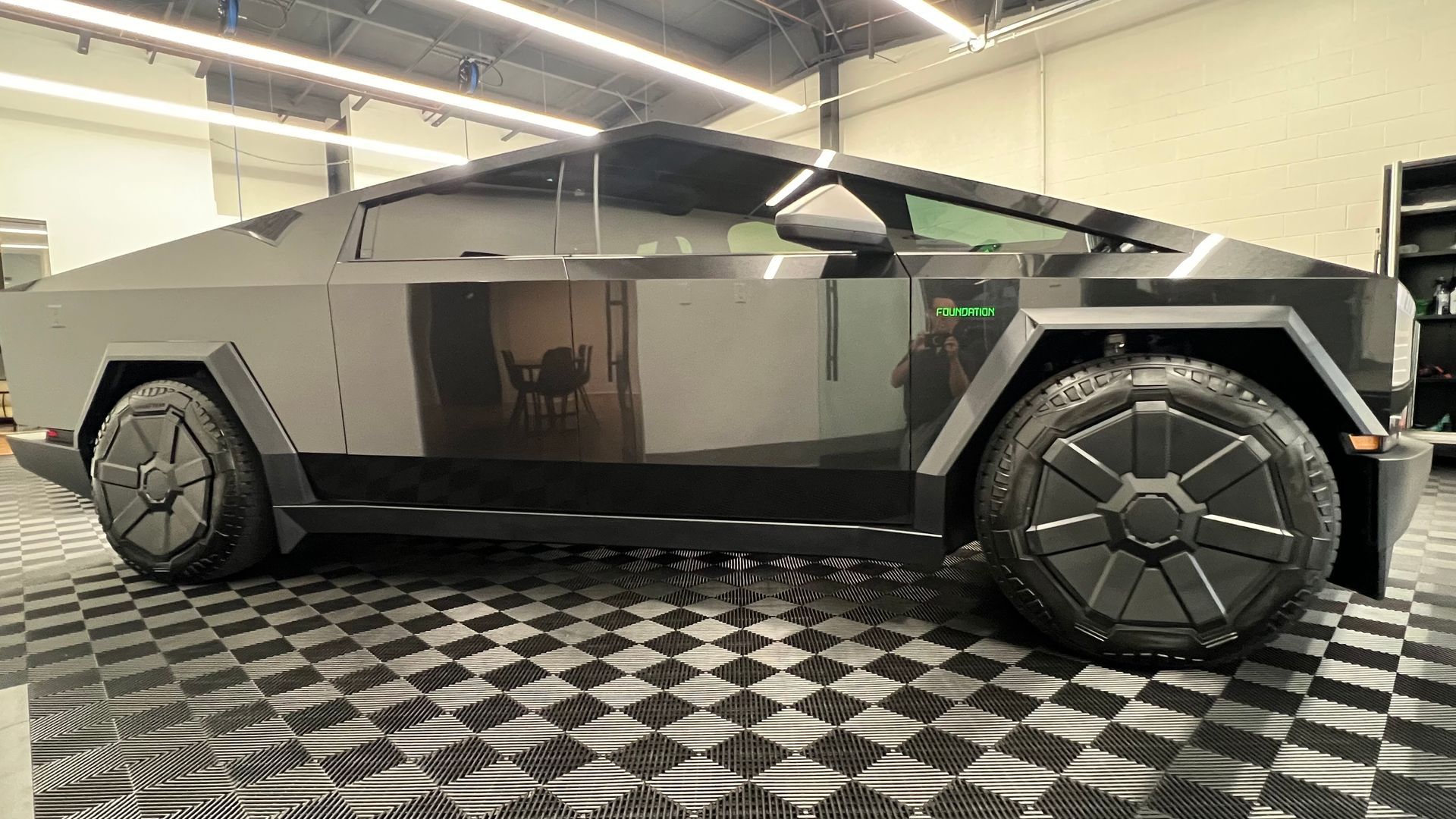 Tesla Cybertruck, a dark gray, angular electric pickup truck with large, black, geometric wheel covers, parked indoors.