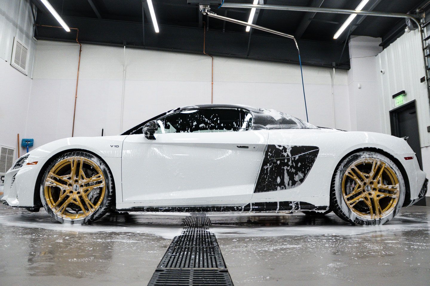 White Audi R8 convertible being washed at a car wash with golden rims, covered in soap. The car is parked indoors.