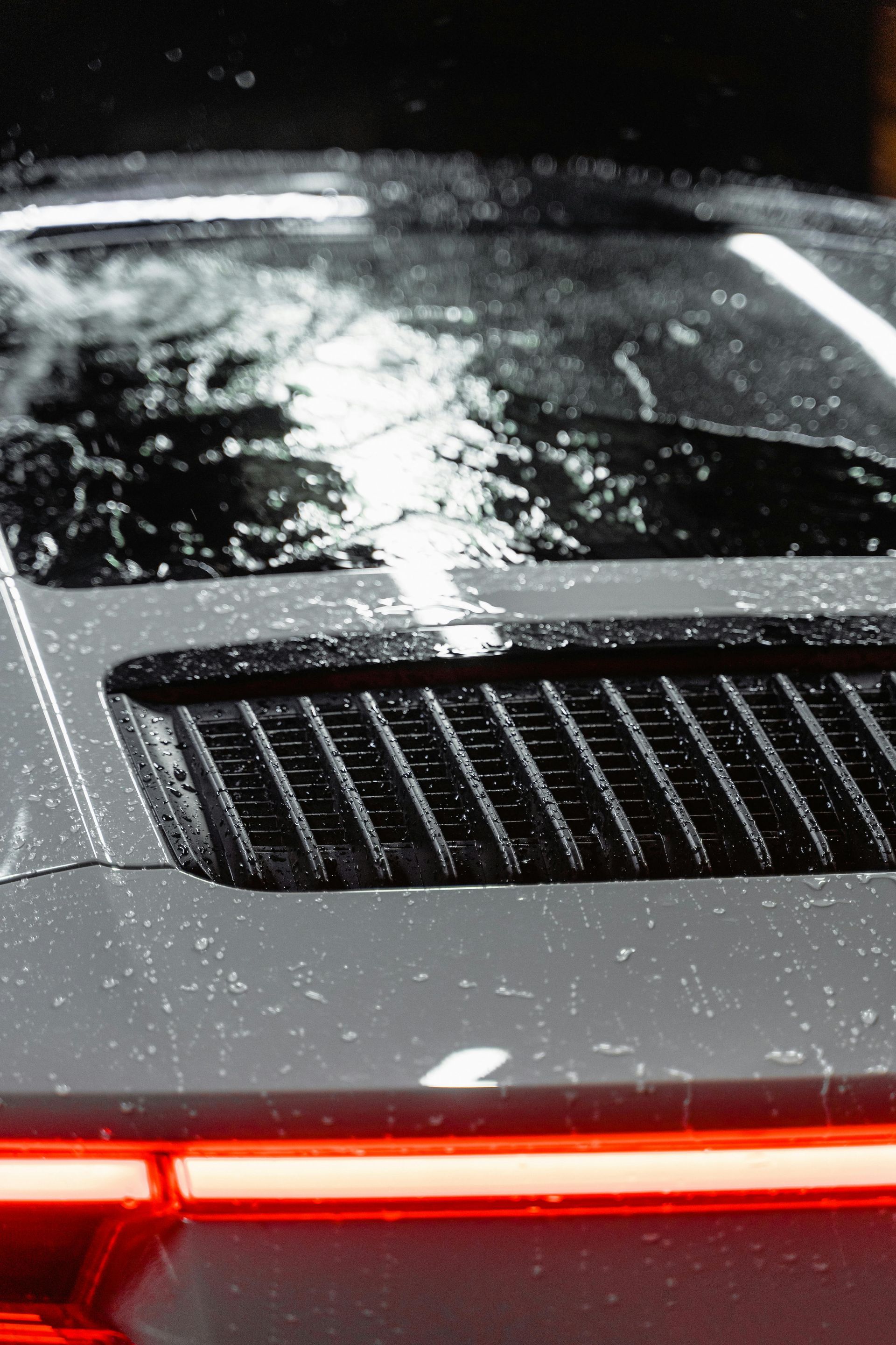 Close-up of a dark grey Porsche car's rear, wet from rain. Horizontal vents sit above the rear lights.