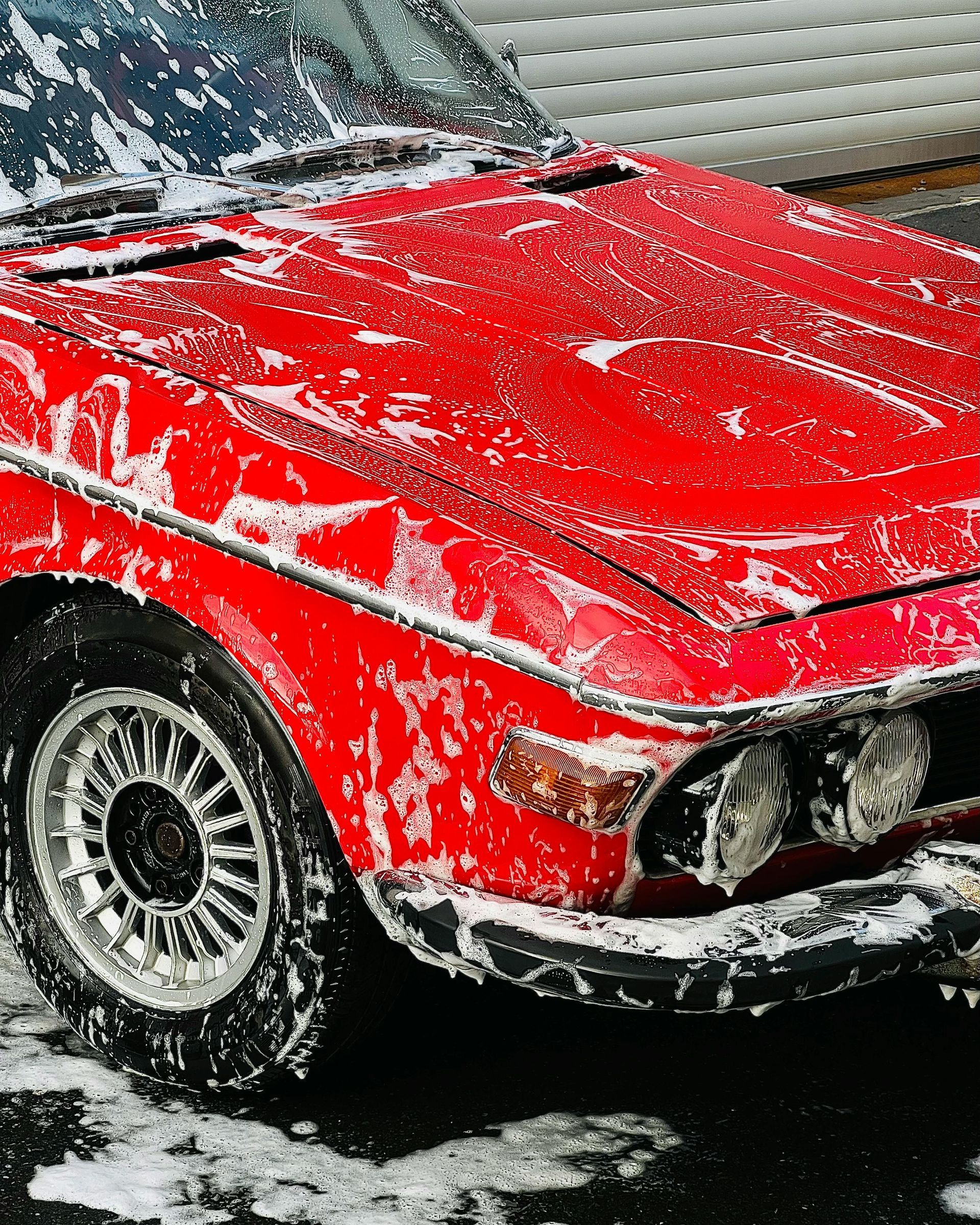 Close-up of a bright green car's front, covered in water droplets. The headlight is lit against a blurred background.
