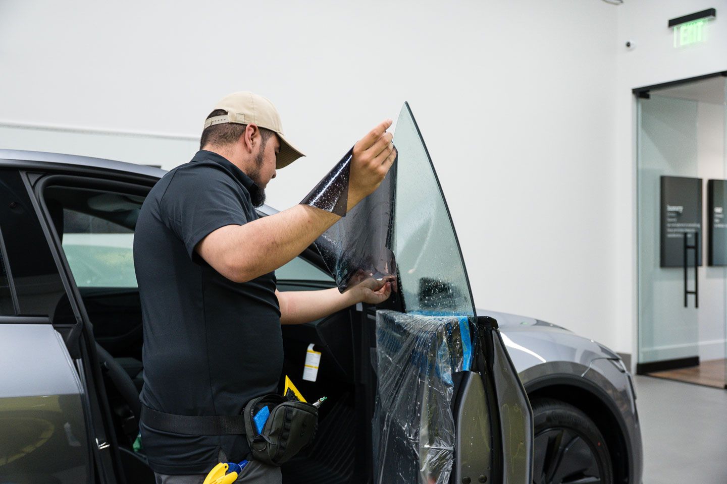 A man installing window tint on a gray car inside a well-lit shop. He holds the tint film, carefully aligning it with the glass.