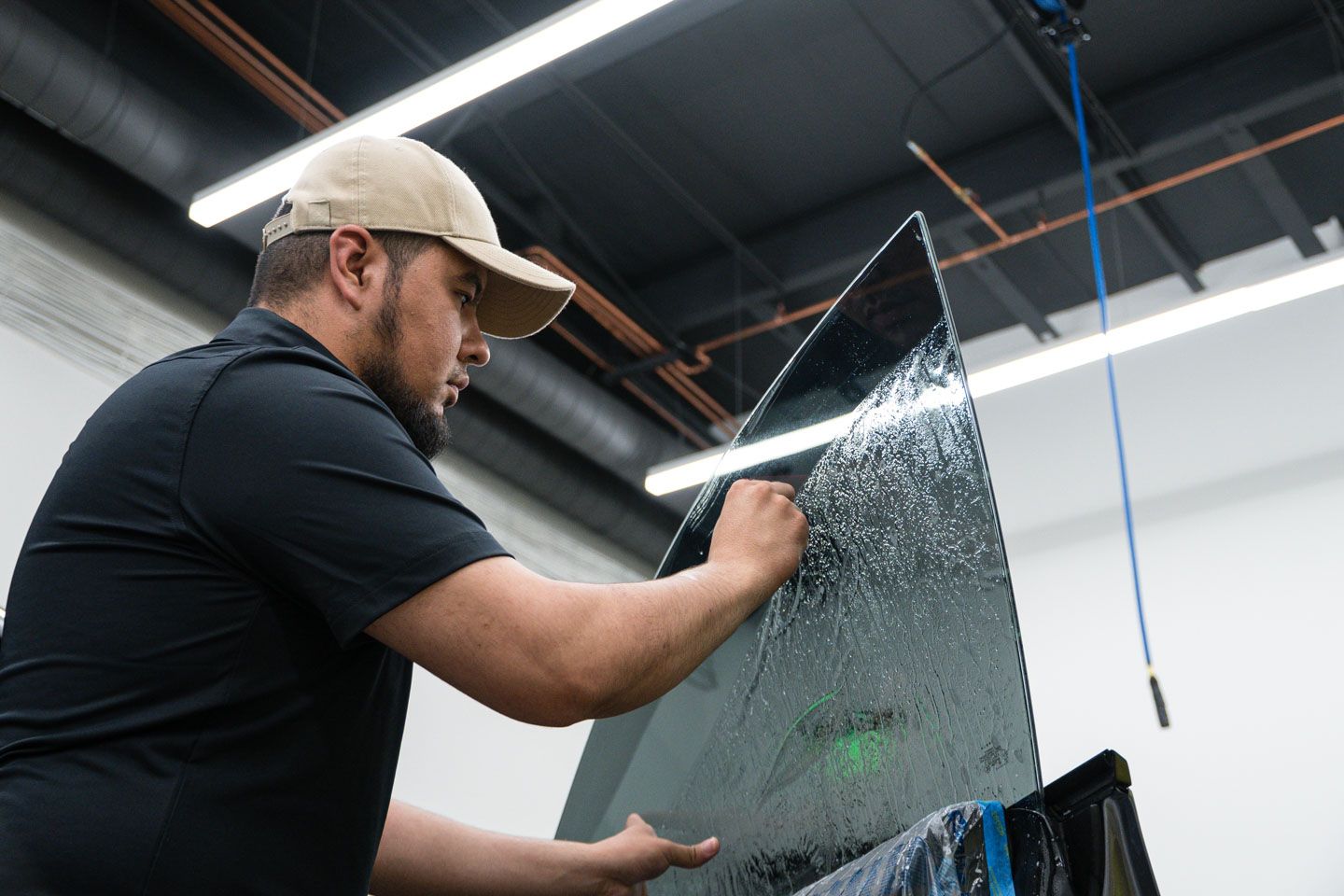 A man in a cap applies tint film to a car window in a workshop, holding the glass up with both hands.