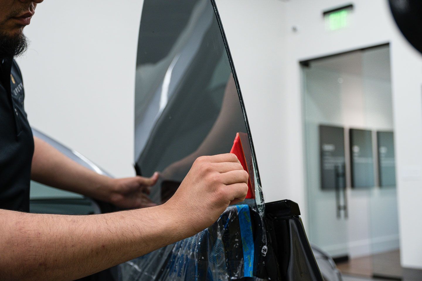 A person applies tint film to a car window using a red squeegee in a bright, modern shop.