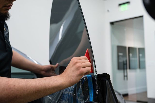 A person applies tint film to a car window using a red squeegee in a bright, modern shop.