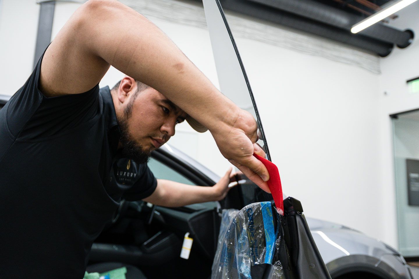 A man is applying window tint to a car window using a red tool. He's in a shop with a gray car.