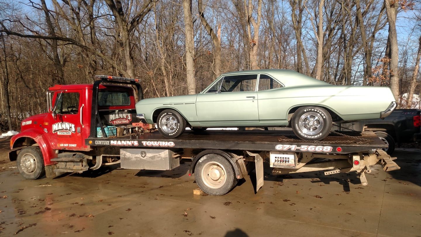 Red tow truck carrying a mint-green classic car on a flatbed in a muddy wooded area