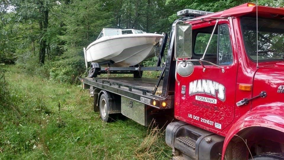 Red tow truck hauling a white boat on a flatbed along a muddy wooded path